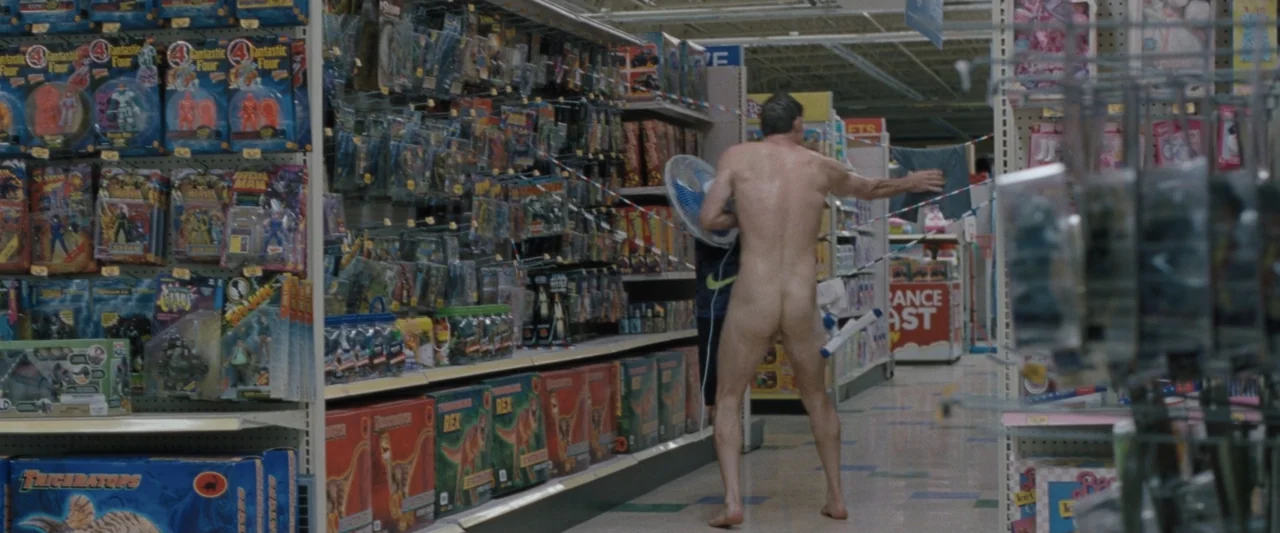 Roofman (2025) movie still: fullbody — A well-lit toy aisle is filled with rows of packaged action figures on shelves. A nude man walks through the…