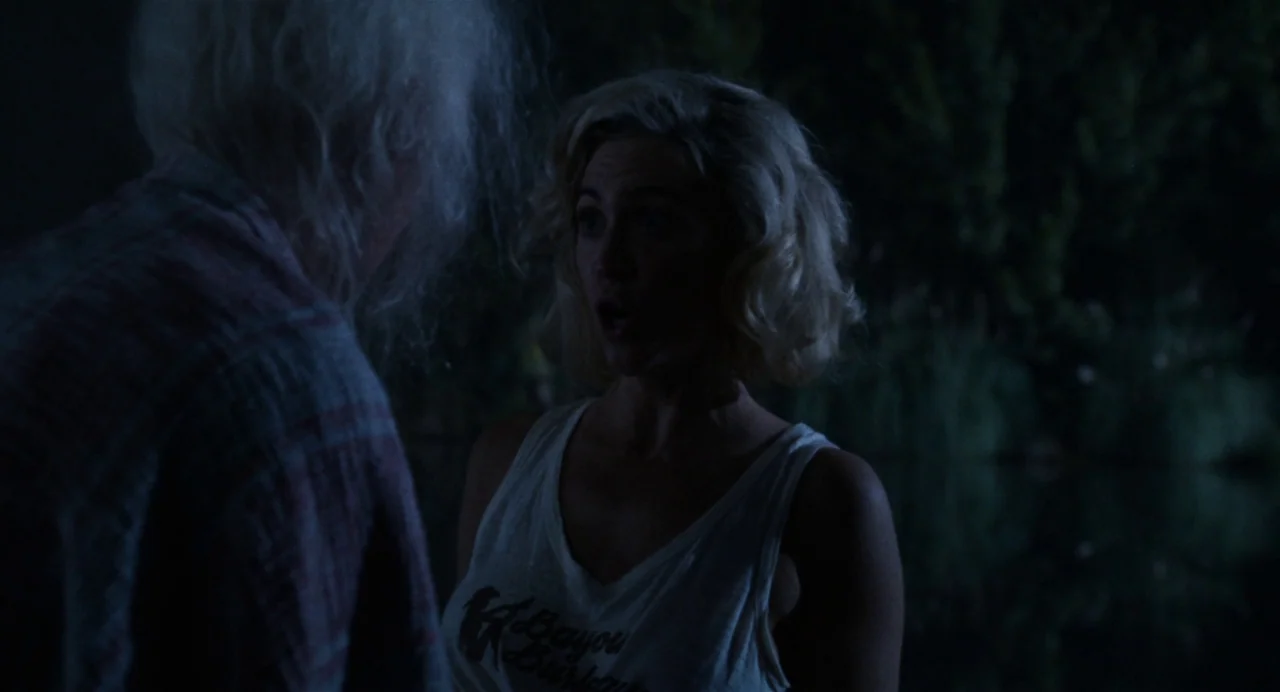 X (2022) movie still: medium — Two individuals stand in a dark, outdoor area at night with dense trees nearby. The woman faces an older person with…