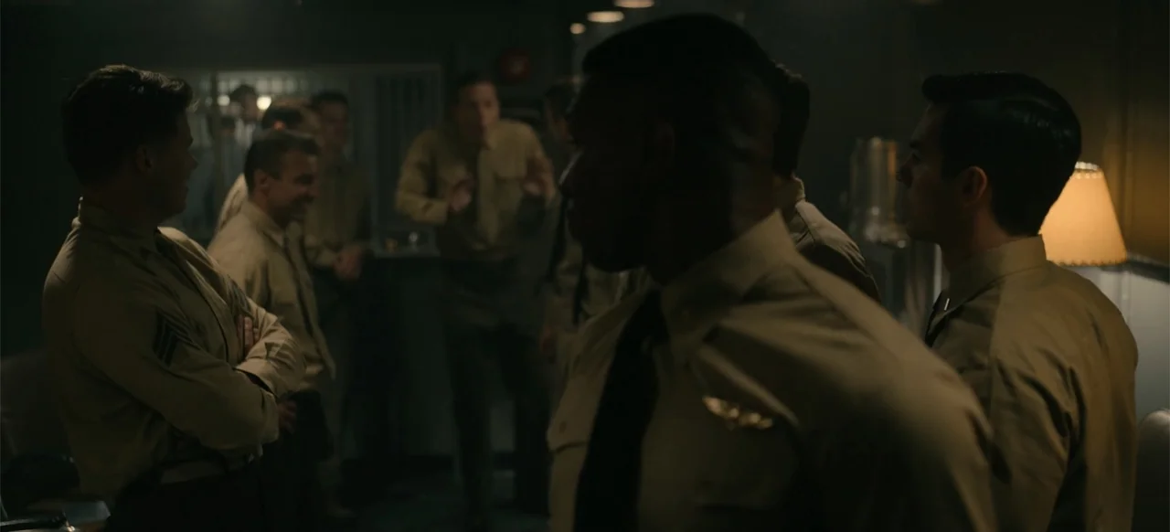 Devotion (2022) movie still: medium — A dimly lit room with several men in tan military uniforms standing together. The men engage in conversation…
