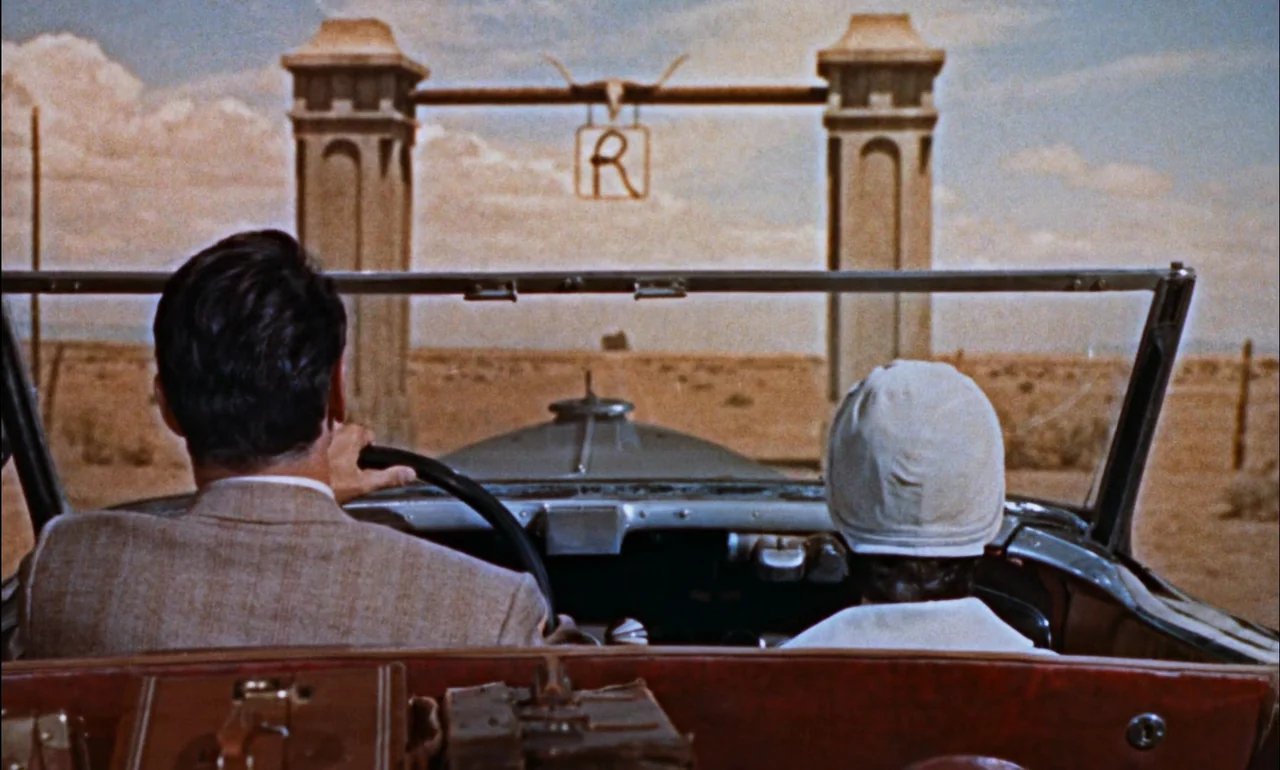 Giant (1956) movie still: medium — A dry desert landscape features a large concrete gate marked with the letter R. A man and a woman in a vintage…