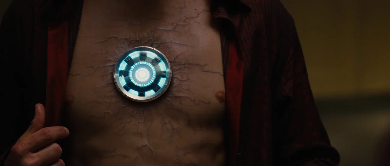 Iron Man 2 (2010) movie still: closeup — A man's torso is shown with a glowing arc reactor embedded in the chest. Dark, vein-like scar tissue…