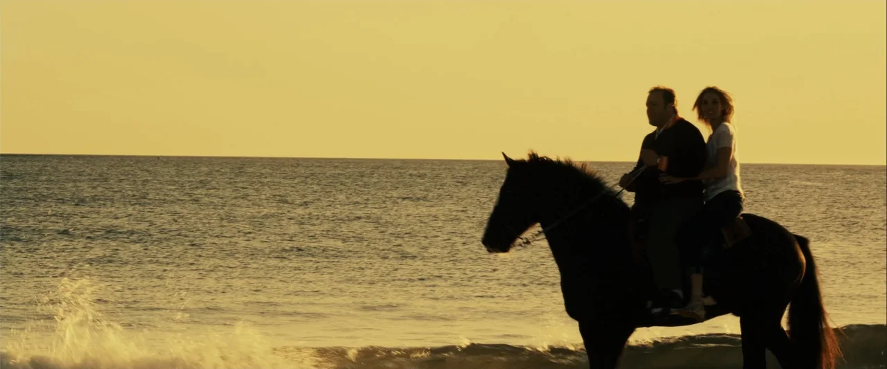 Zookeeper (2011) movie still: fullbody — A man and woman ride a horse through the surf at sunset. The ocean horizon meets a clear sky while waves…