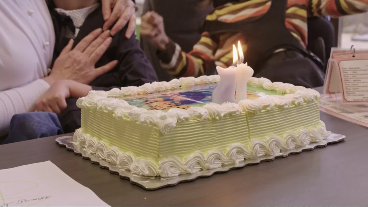 Finding the Way Home (1991) movie still: medium — A rectangular birthday cake with white frosting and two lit candles sits on a table. Two…