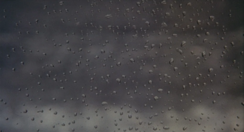 The Go-Between (1971) movie still: closeup — A clear glass pane is covered in various sized water droplets. The background consists of an…