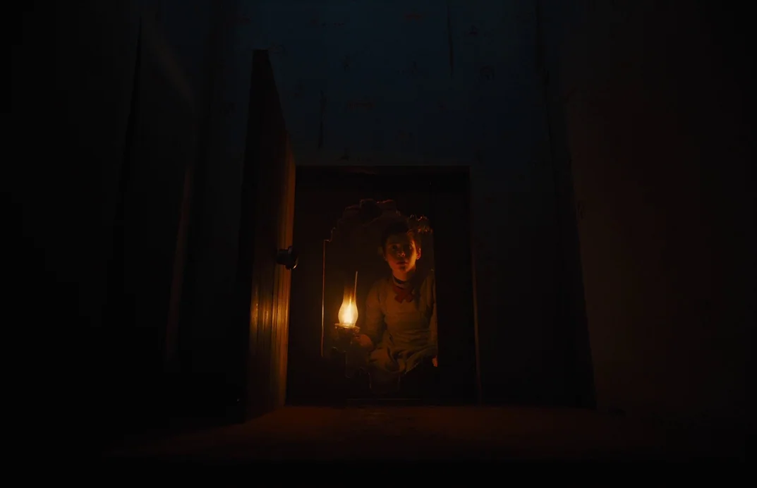 Gretel & Hansel (2020) movie still: medium — A dimly lit, cramped space behind a wall features a small jagged opening. A young boy peeks through the…
