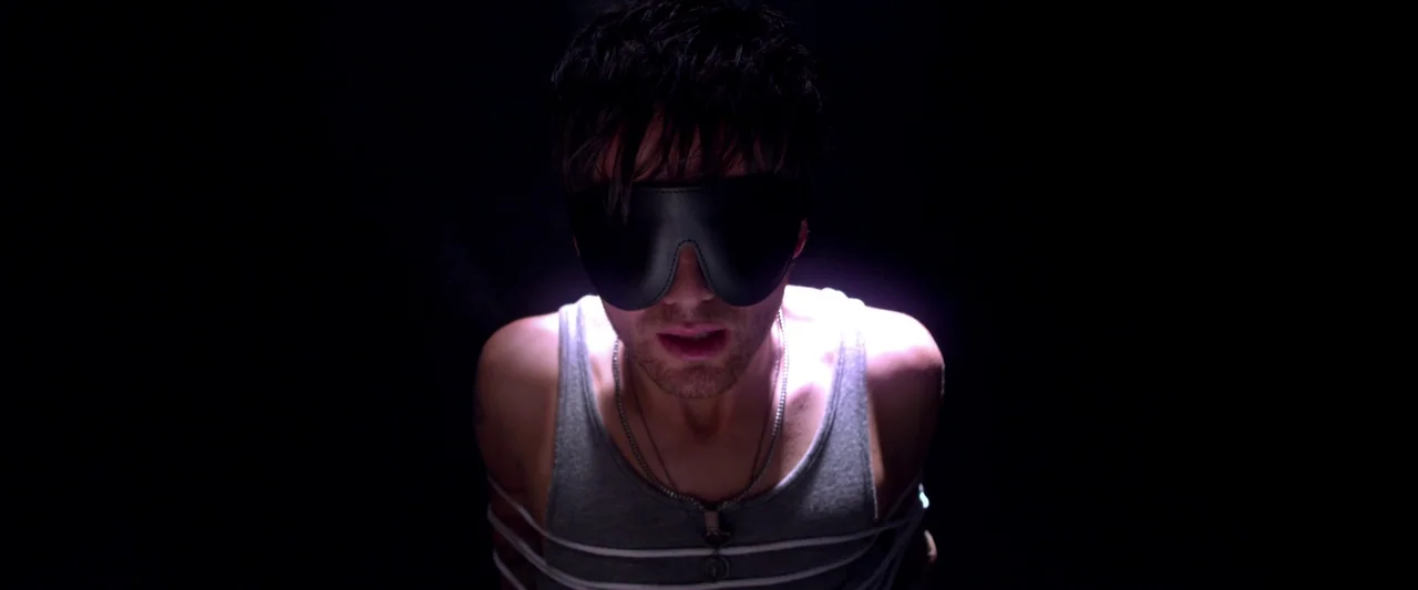 Kaboom (2010) movie still: medium — A dark, featureless environment surrounds a man tied with white rope. The man wears a leather blindfold while…