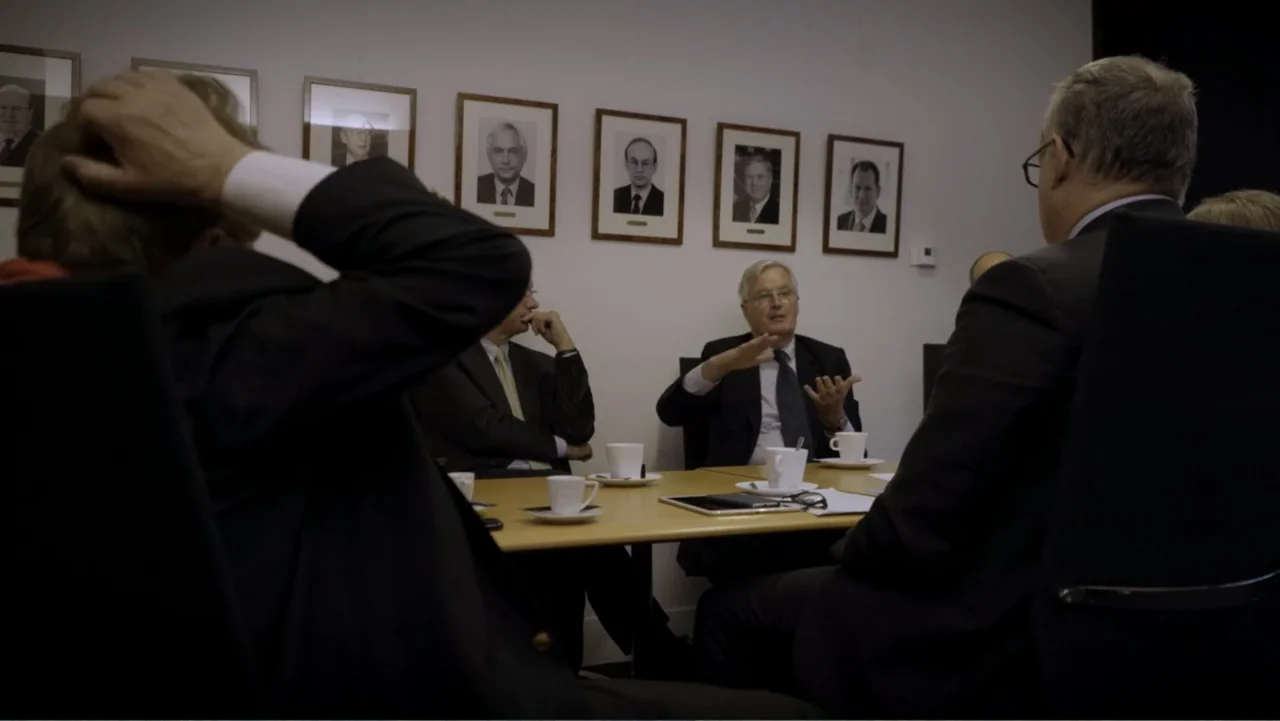 Brexit: Behind Closed Doors (2019) movie still: medium — A conference room with framed portraits lining the white wall above a wooden table. A man…