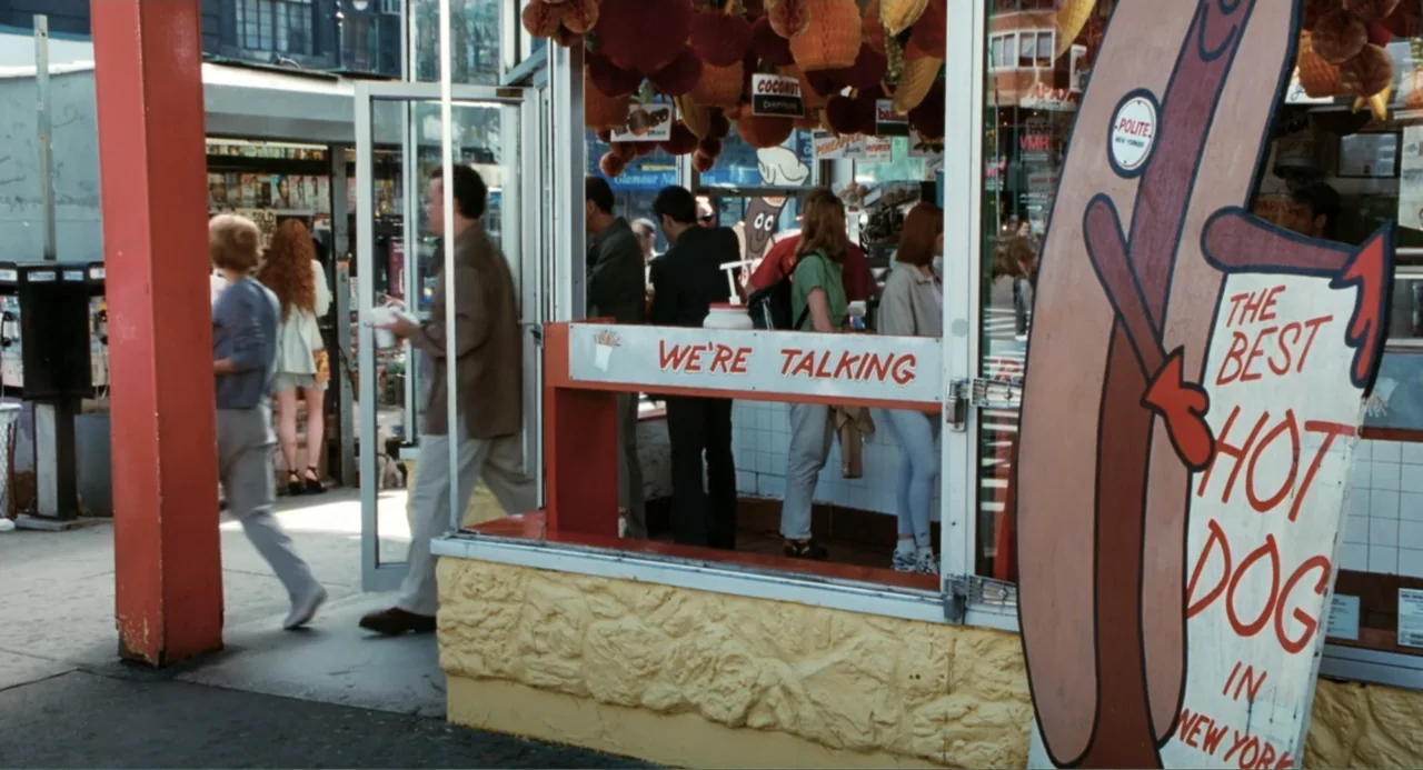 You've Got Mail (1998) movie still: medium — A bustling New York hot dog stand with a prominent painted sign on the right. A man in a tan shirt…