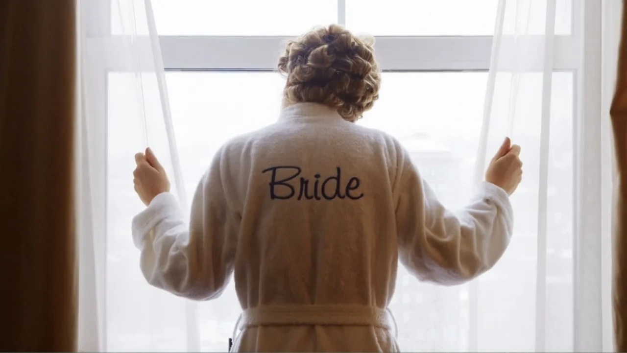 Suited (2016) movie still: medium — A hotel room features a large window draped with white curtains. A woman wearing a white 'Bride' robe stands…