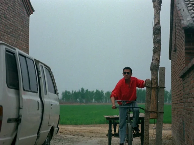 Black Snow (1990) movie still: fullbody — A rural dirt path bordered by red brick walls and a green field. A man in a red shirt sits on a tricycle…