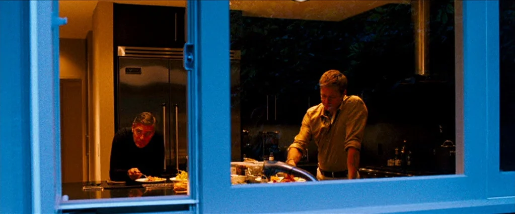 Ocean’s Thirteen (2007) movie still: wide — A dark modern interior visible through a window frame at night. One man eats while the other man stands…