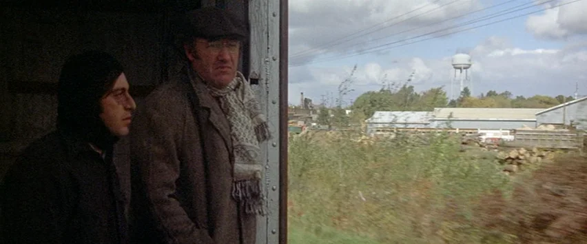 Scarecrow (1973) movie still: medium — Two men stand inside the open side door of a cargo train car. Outside, a rural landscape with industrial…