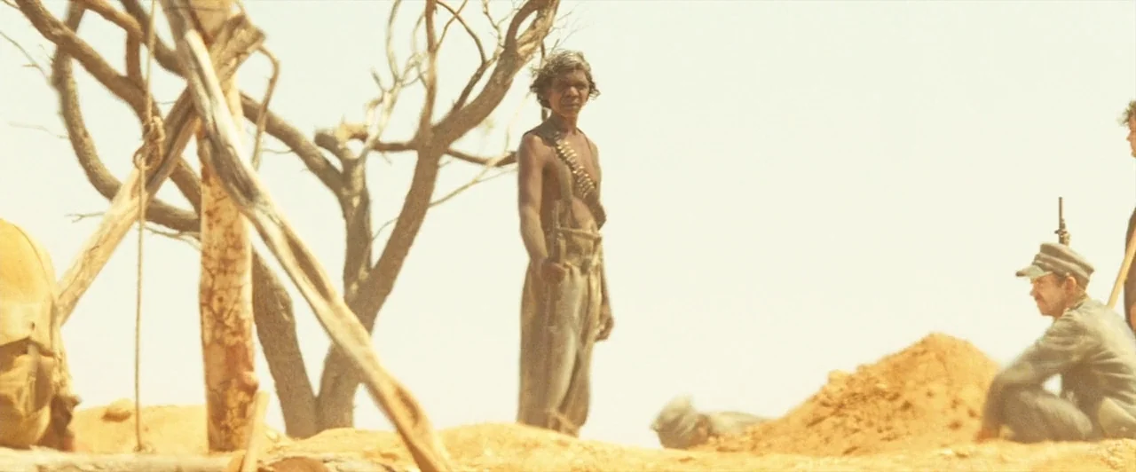 The Proposition (2005) movie still: wide — A dry desert landscape features sparse dead trees and piles of loose sandy earth. An indigenous man…