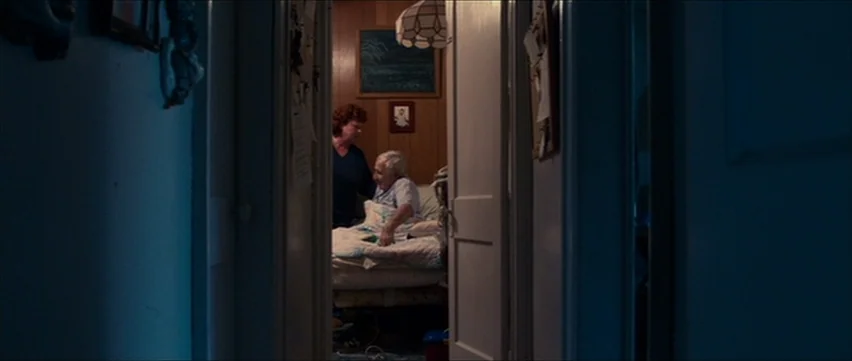 Bubble (2005) movie still: wide — A dark hallway leads into a small illuminated room with wood-paneled walls. A woman in a blue shirt stands by a…