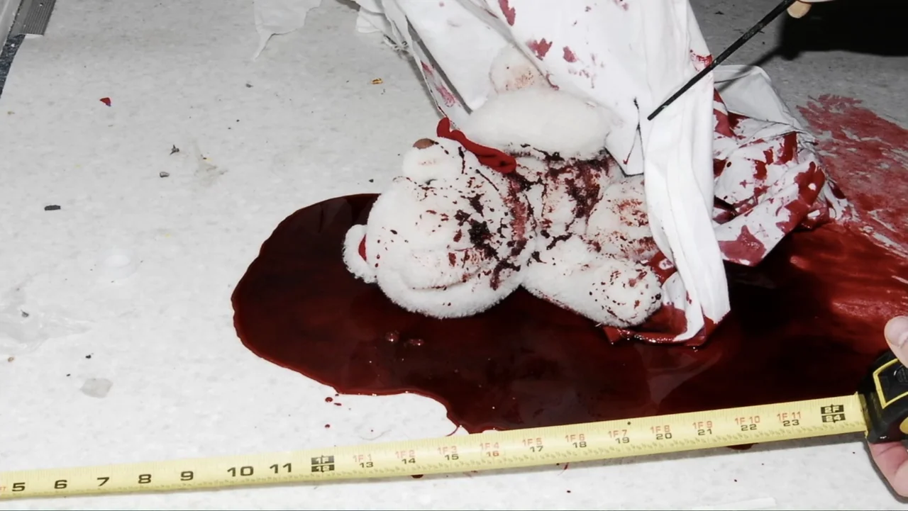 Savageland (2015) movie still: closeup — A white tile floor is covered with a large pool of dark red blood. A soiled white teddy bear rests in the…