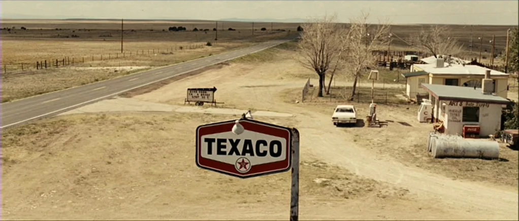 No Country For Old Men movie still: establishing — A desolate desert gas station stands beside a long, empty paved road. A red and white Texaco sign…