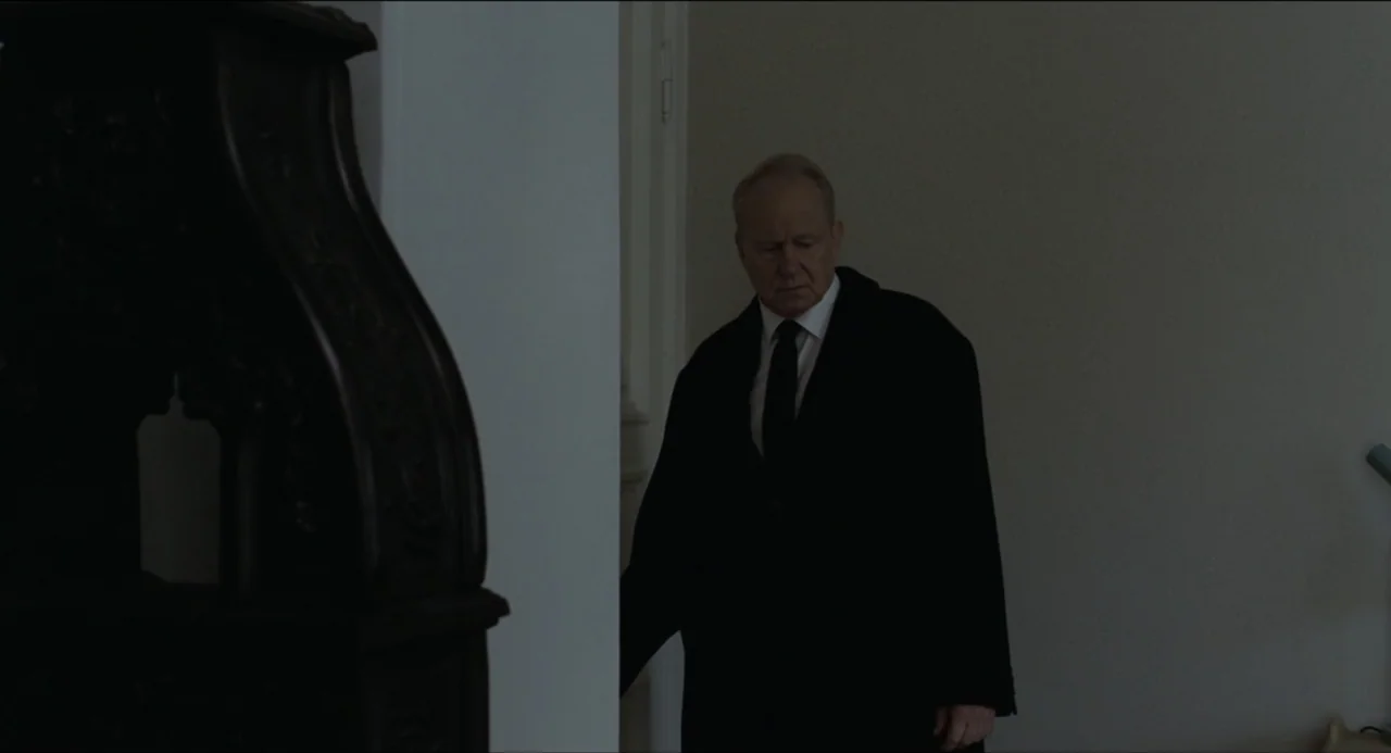 Sentimental Value (2025) movie still: medium — An interior hallway features a white door frame and a blurred dark antique object. An older man in a…