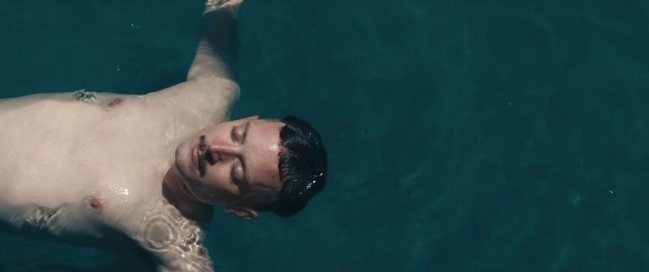 Come To Daddy (2019) movie still: medium — A man floats on his back in dark blue water. The surface ripples around his body as he maintains a…