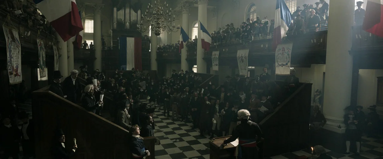 Napoleon (2023) movie still: wide — A historic wooden interior features a checkered stone floor and a elevated speaking podium. A large crowd of men…