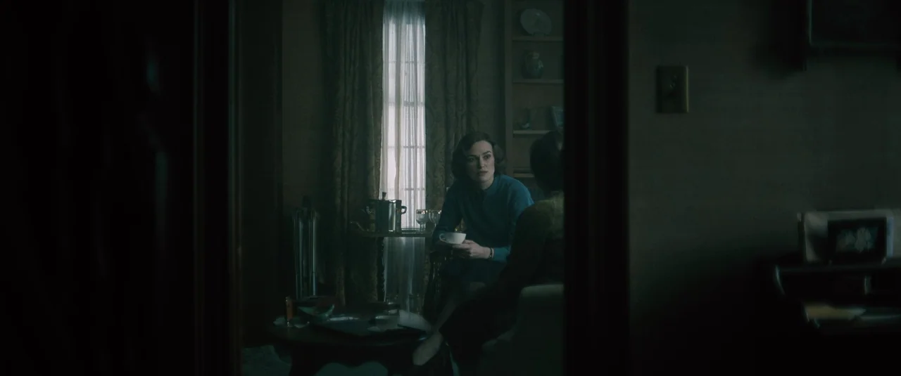 Boston Strangler (2023) movie still: medium — A dimly lit room features a woman seated by a window holding a cup. The view is framed through a dark…