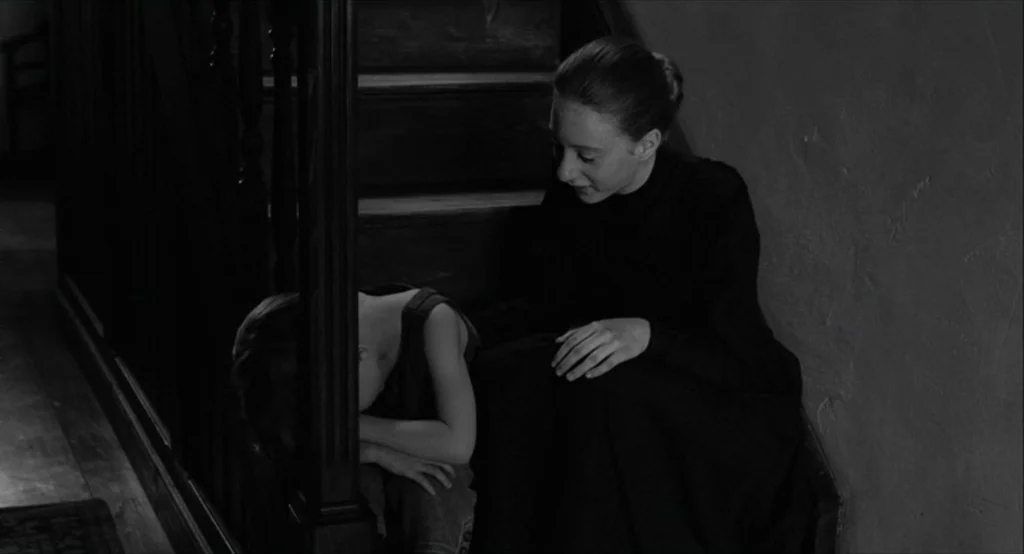 The White Ribbon (2009) movie still: medium — A wooden staircase in a dimly lit home with a dark banister and railings. A woman in a black dress…