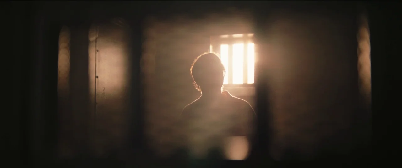 The Batman (2022) movie still: medium — A dimly lit room features a central barred window letting in intense daylight. A person stands silhouetted…