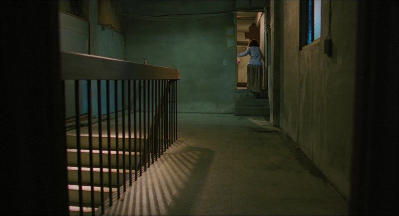 Goodbye, Dragon Inn (2003) movie still: wide — A dimly lit concrete stairwell landing features a dark metal railing on the left. A woman with…