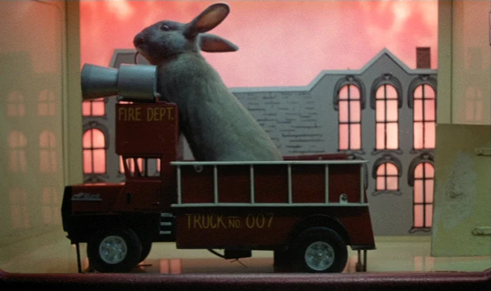 Stroszek (1977) movie still: medium — A grey rabbit sits inside the bed of a small red toy fire truck. The scene is staged in front of a flat,…