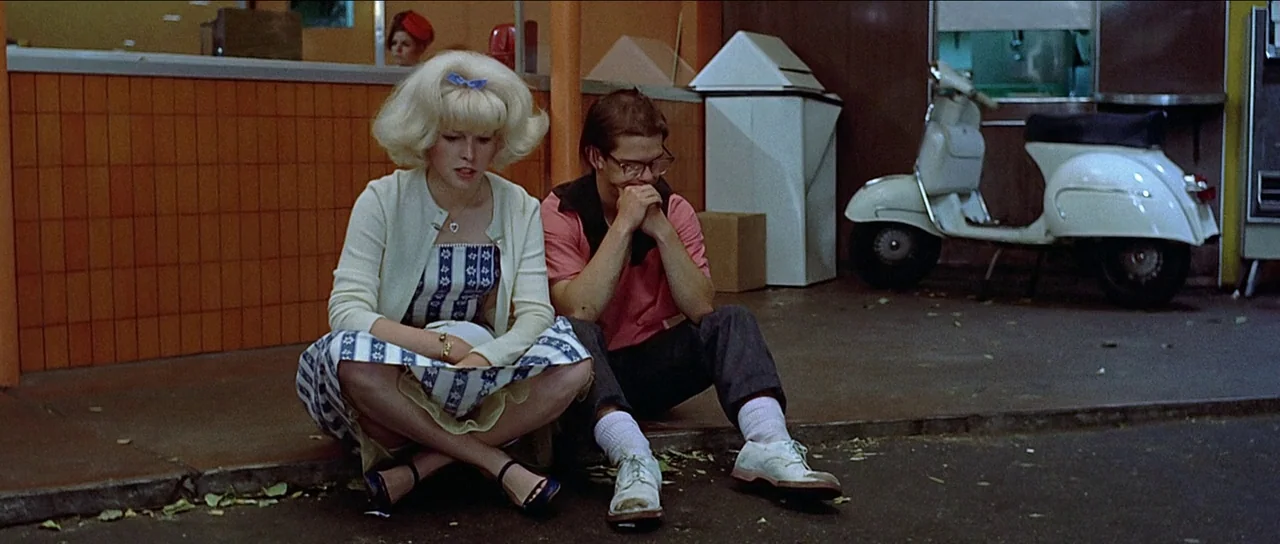 American Graffiti (1973) movie still: fullbody — A pair of young adults sit on the pavement outside a tiled commercial building. A white vintage…
