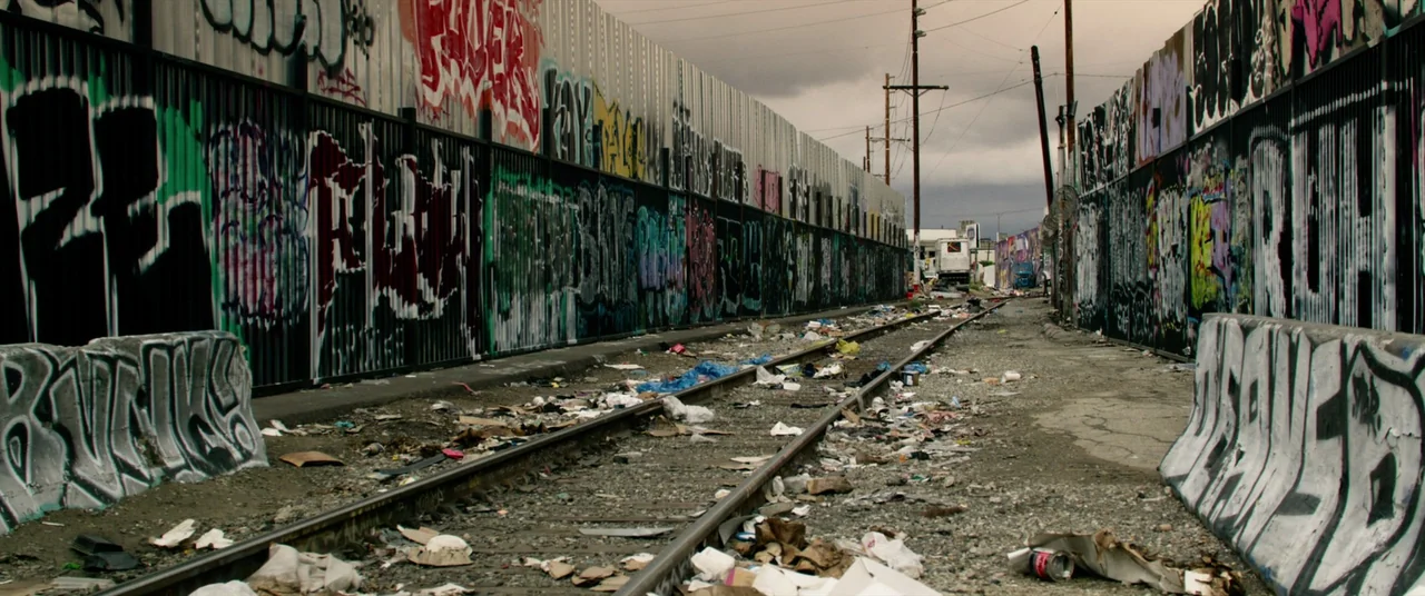 Ambulance (2022) movie still: wide — Railway tracks run through a narrow corridor flanked by high walls covered in graffiti. Litter and debris are…