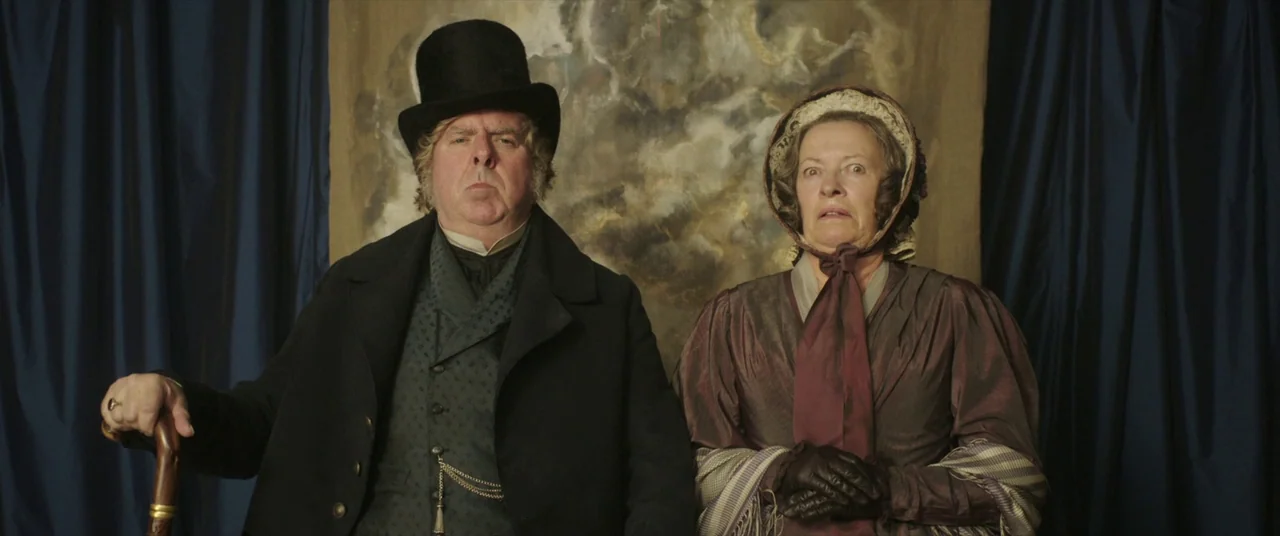 Mr. Turner (2014) movie still: medium — A man and woman pose formally in front of blue curtains and a backdrop. The subjects stand side by side…