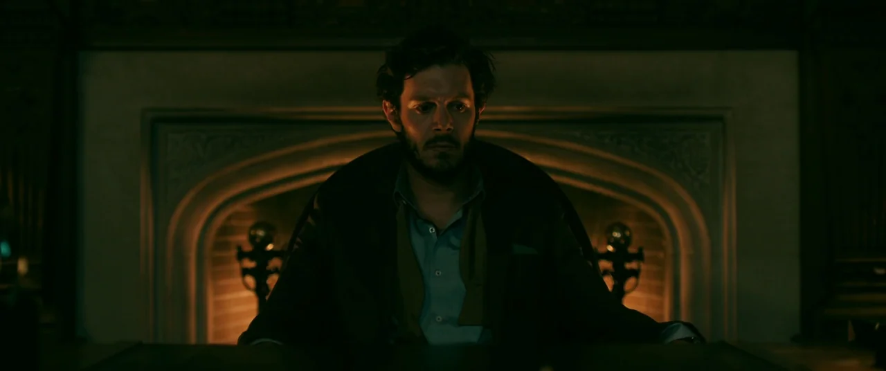 Ready or Not (2019) movie still: medium — A dark study features an ornate fireplace glowing with low embers in the background. A man sits at a desk…