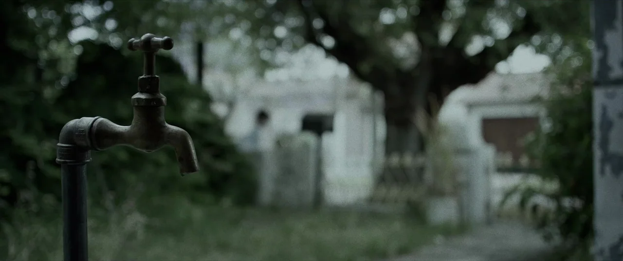 Terrified (2017) movie still: closeup — A metal spigot stands in the foreground against an out-of-focus yard. A house and trees are visible in the…