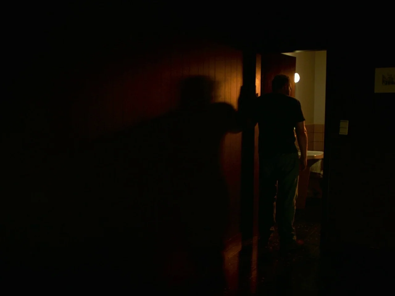 Violet (2014) movie still: fullbody — A dark corridor leads into a brightly lit washroom with tiled walls. A man in dark clothes stands in the…