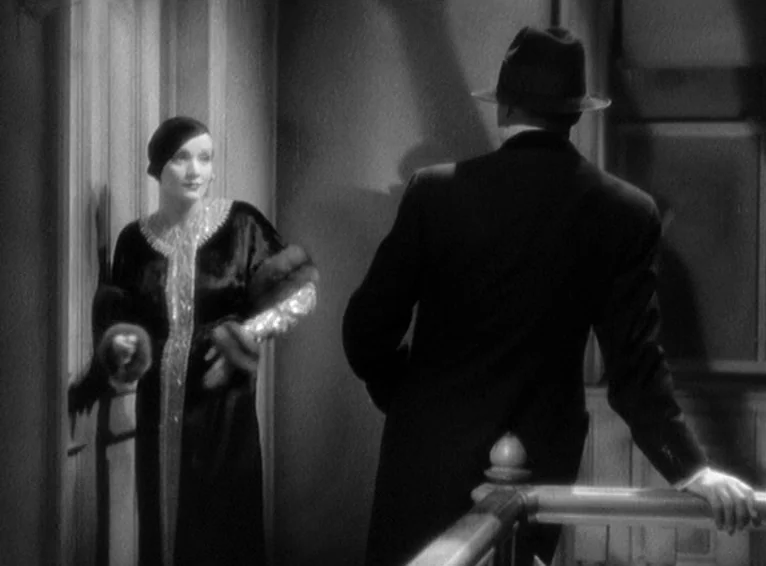 Blonde Venus (1932) movie still: medium — A dimly lit staircase area features a man in a hat and a woman. The woman in a gown stands by an open door…