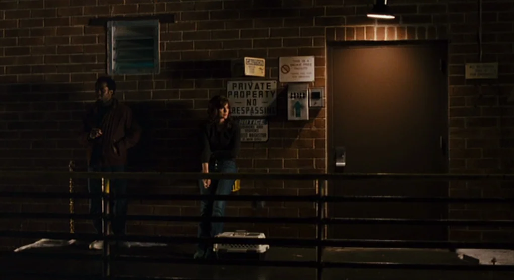 The Savages (2007) movie still: wide — A brick wall at night features multiple signage placards and a metal access door. Two people stand near a…