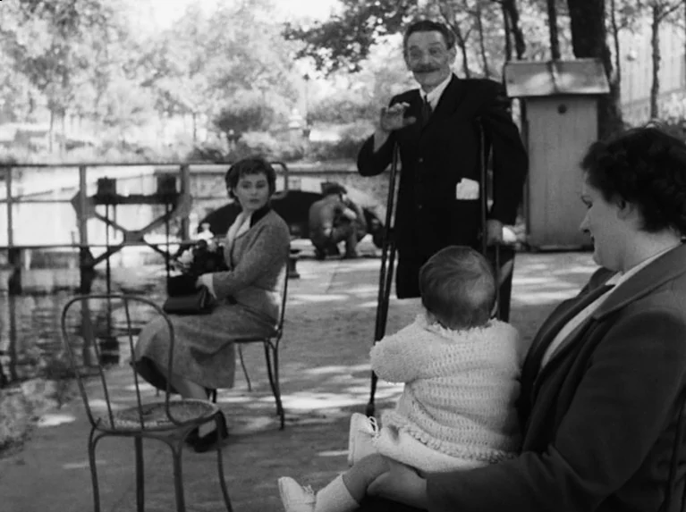 Waiting Women (1952) movie still: medium — A park pathway by a waterway features several adults and a small infant. The man stands on crutches while…