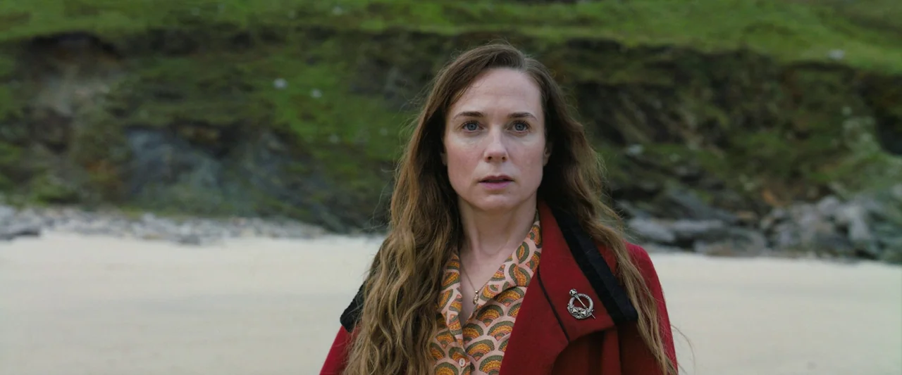 The Banshees of Inisherin (2022) movie still: medium — A woman stands on a sandy beach in front of a rocky grassy cliff. She gazes ahead with a…