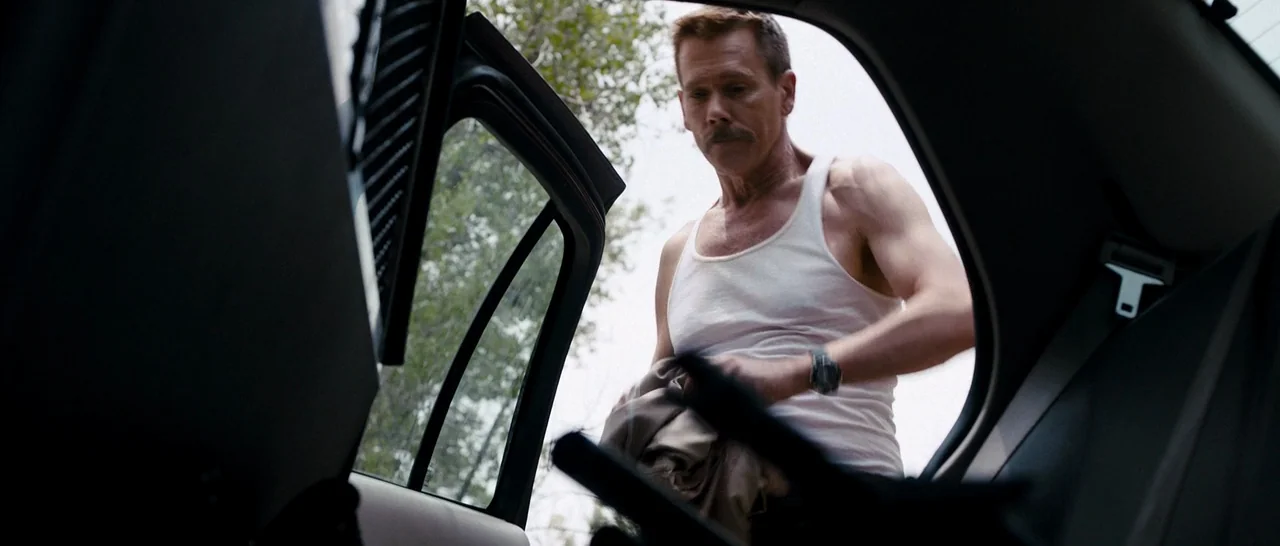 Cop Car (2015) movie still: medium — The interior of a vehicle seen from the perspective of the backseat. A middle-aged man in a white tank top…