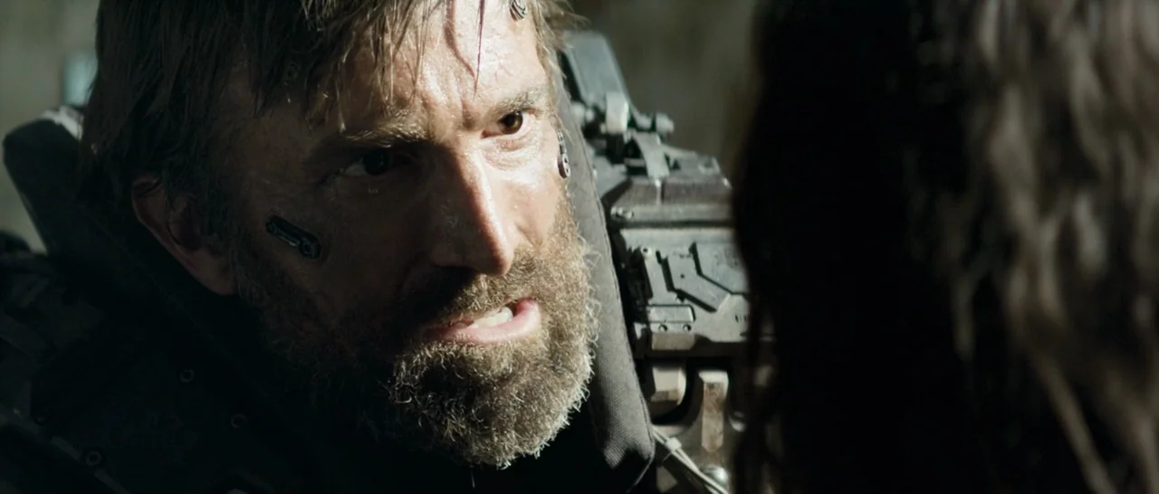 Elysium (2013) movie still: closeup — A close view of a man wearing military gear in a tactical environment. The man makes eye contact with another…