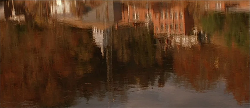 All The Real Girls (2003) movie still: establishing — A body of water captures the distorted reflection of brick buildings and trees. The water…