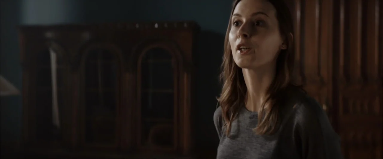 La herencia (2010) movie still: medium — A dimly lit interior space features a dark wooden cabinet in the background. A woman in a grey shirt is…