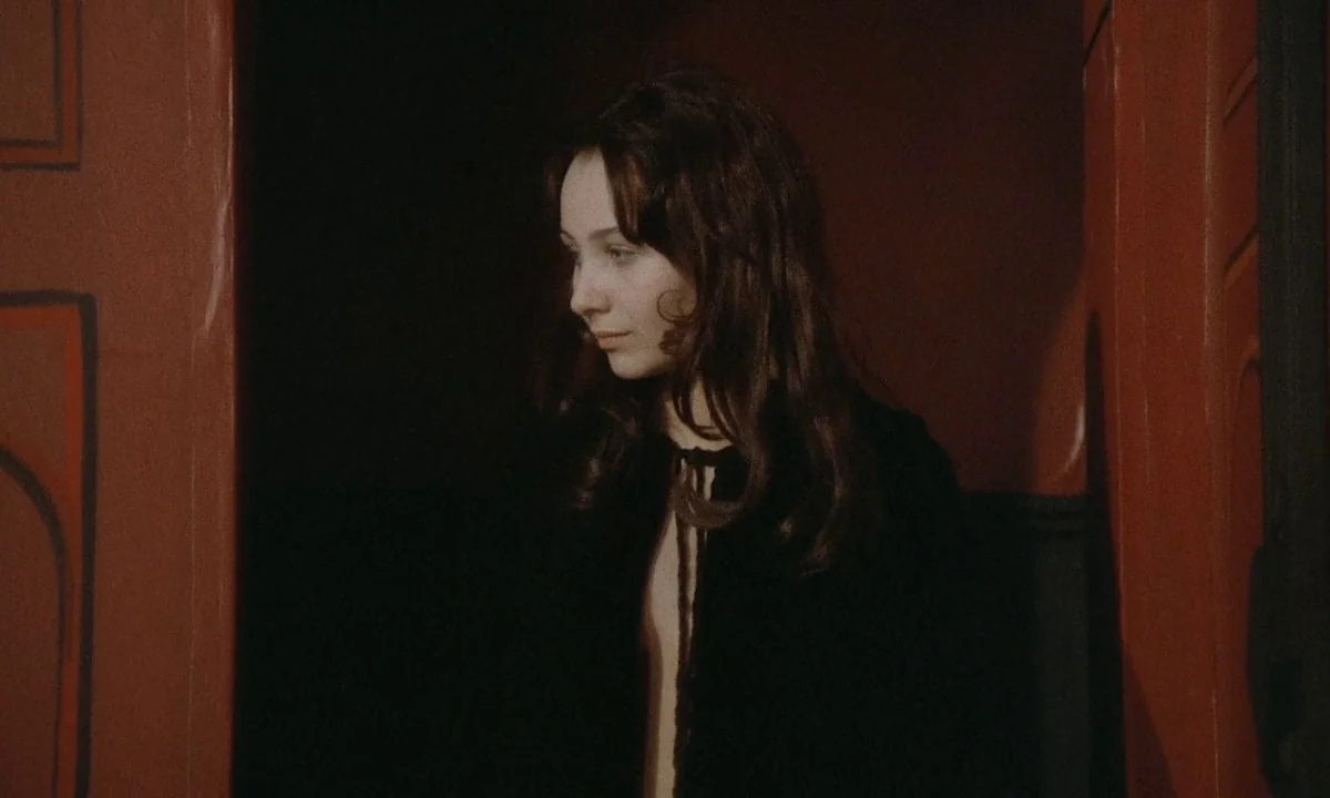 Immoral Tales (1974) movie still: medium — A dark space framed by deep red walls on either side of the frame. A young woman with long dark hair…