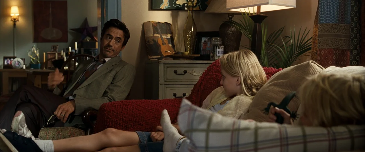 Due Date (2010) movie still: medium — A furnished living area contains a red sofa and wooden furniture with lamps. A man in a suit looks toward two…