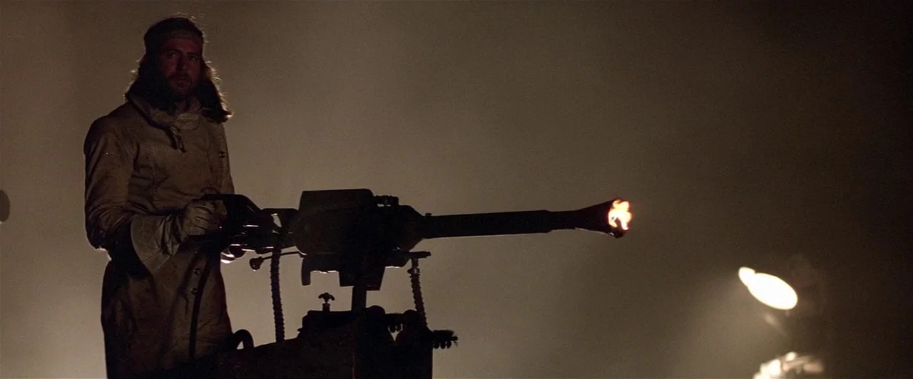 Mad Max 2 (1981) movie still: medium — A man stands in a dark, haze-filled room operating a mounted heavy machine gun. Muzzle fire erupts from the…