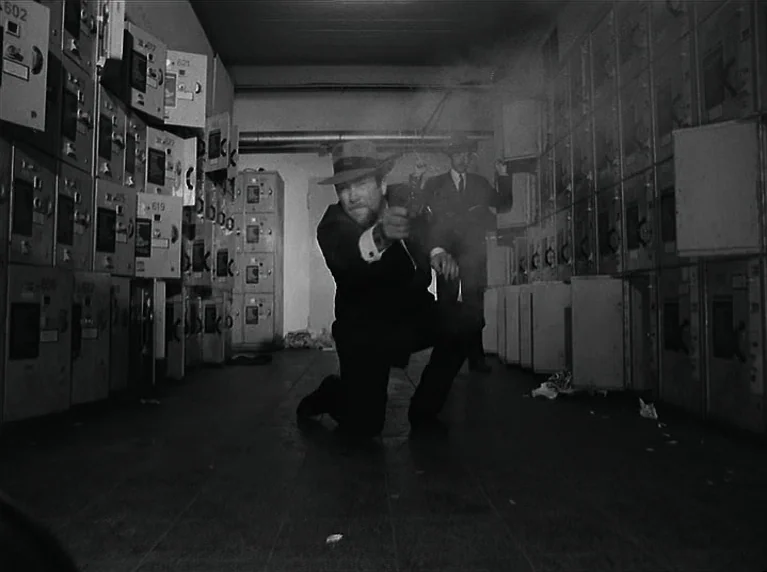 The American Soldier (1970) movie still: fullbody — A dark locker room lined with rows of metallic storage units. A man in a suit kneels on the…