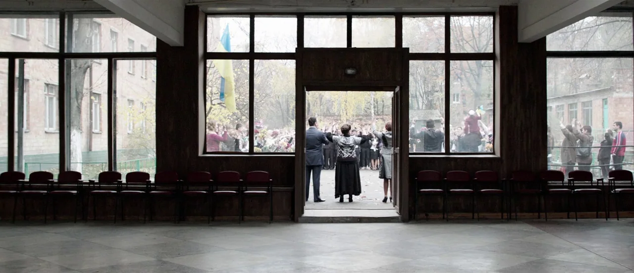 The Tribe (2014) movie still: wide — A tiled interior hall with rows of dark chairs faces a large glass partition. Three adults stand in an open…