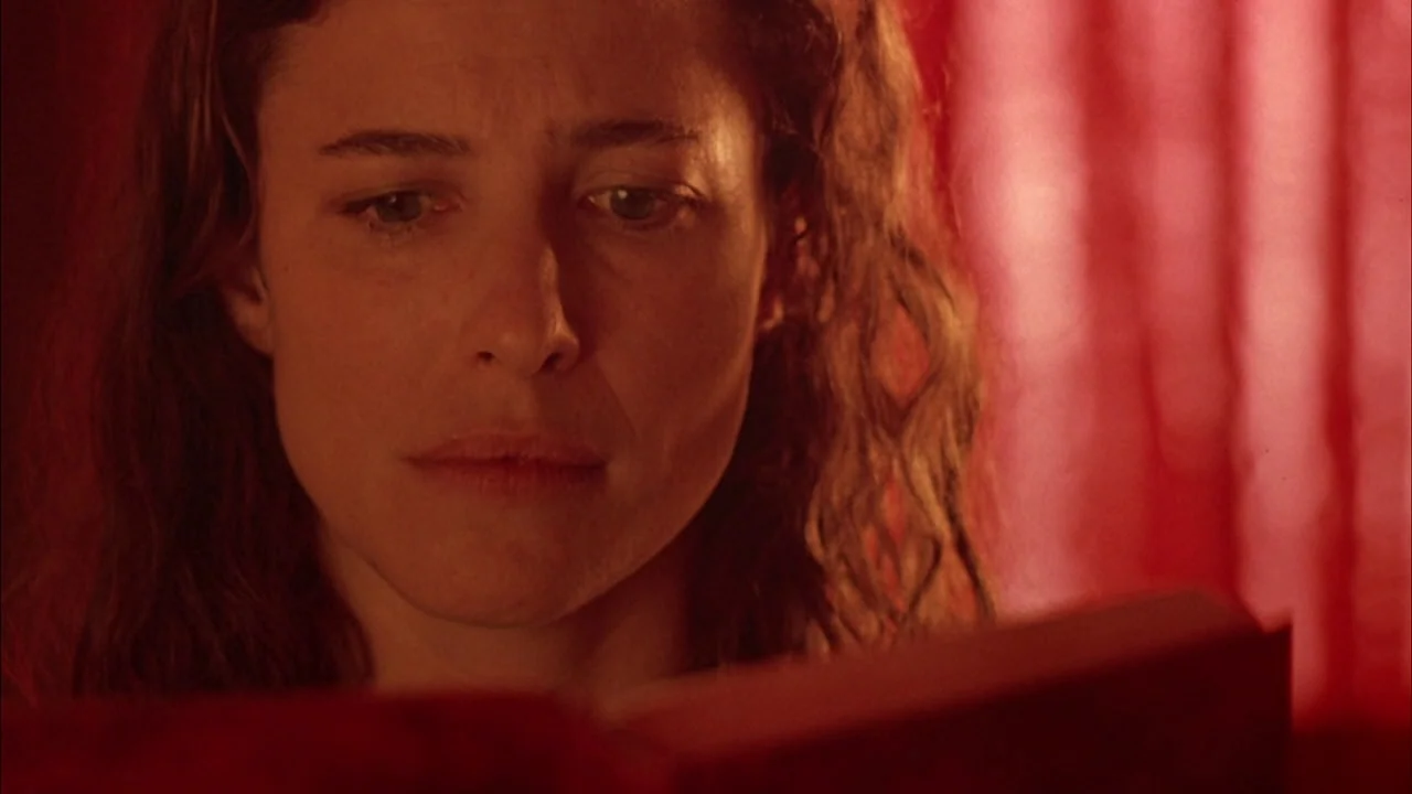 The Rapture (1991) movie still: closeup — A woman reads a book in a dimly lit room with red curtains. The soft light highlights her features against…