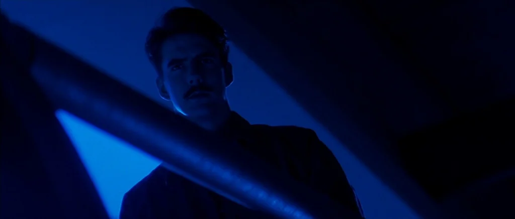The Underneath (1995) movie still: medium — A man stands behind a diagonal metal railing in a dimly lit, blue-hued stairwell. He looks forward while…