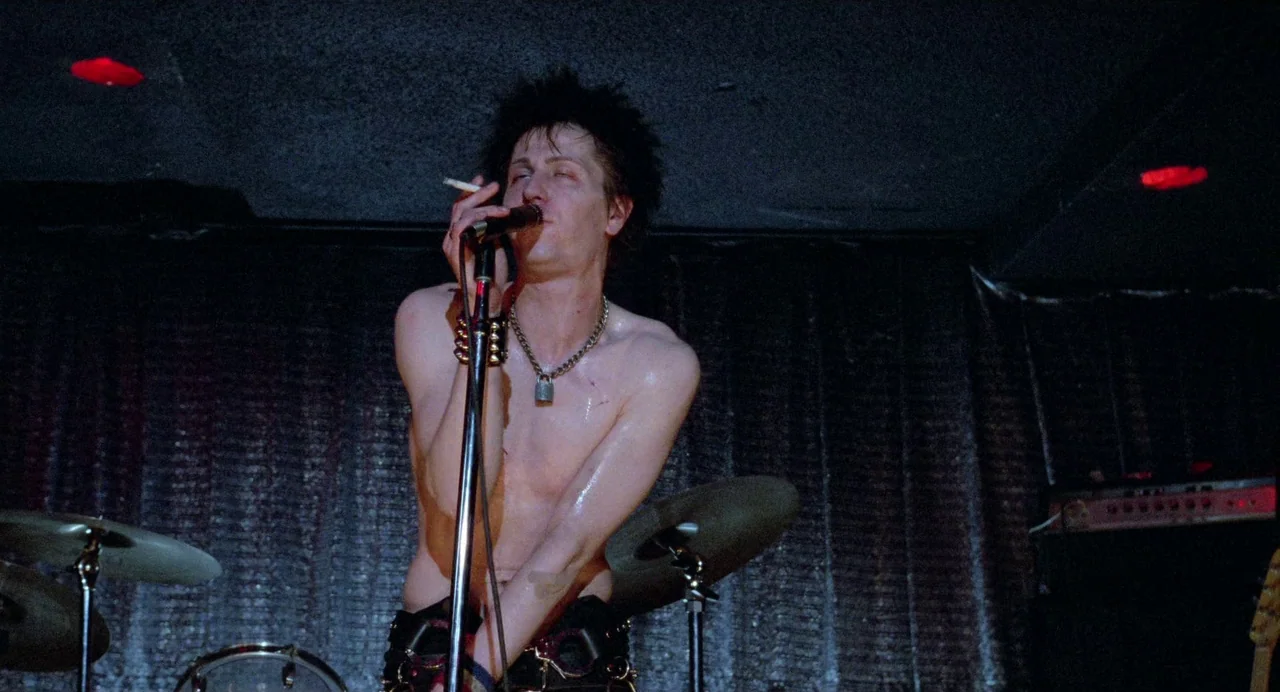 Sid and Nancy (1986) movie still: medium — A dark concert venue with black curtains and musical equipment in the background. A shirtless man with…